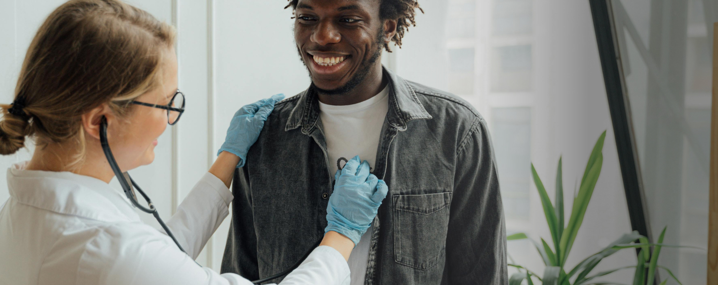 A healthcare professional in a white coat and gloves is using a stethoscope to check the heart or lungs of a smiling young man wearing a denim jacket in a clinical setting.