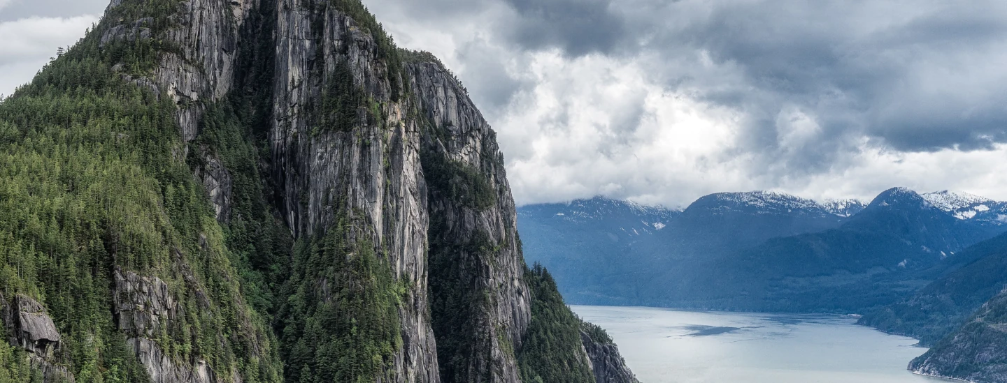 The image captures the view in Squamish BC, a forest-covered cliffs beside a tranquil fjord under a cloudy sky.