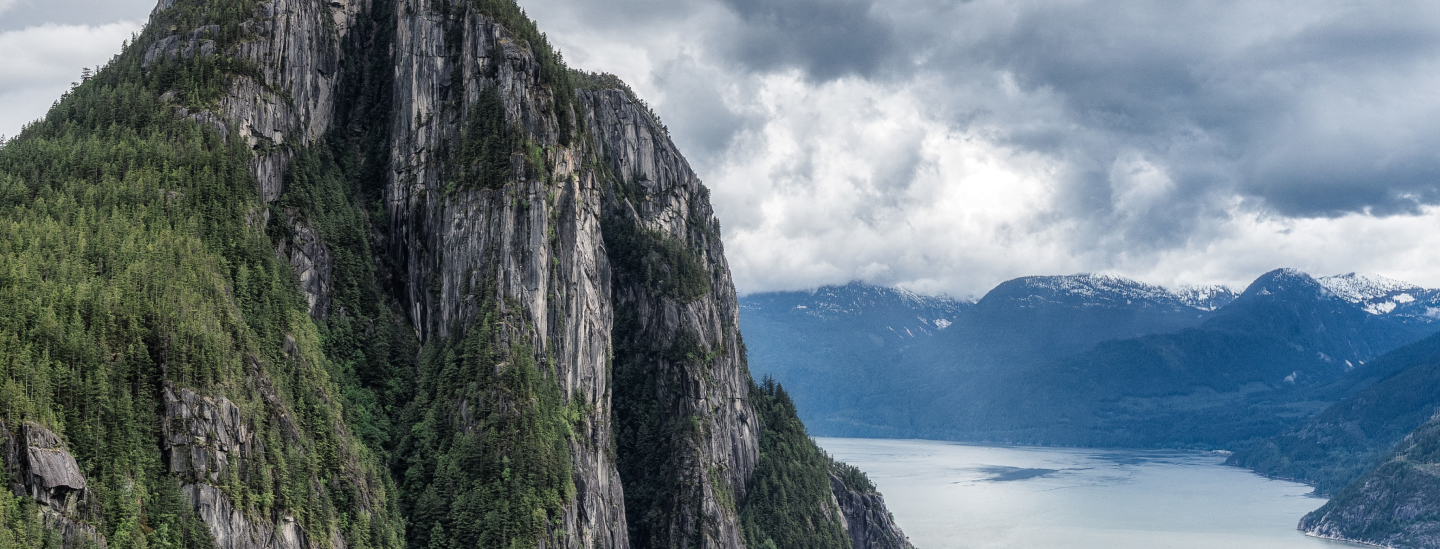 The image captures the view in Squamish BC, a forest-covered cliffs beside a tranquil fjord under a cloudy sky.